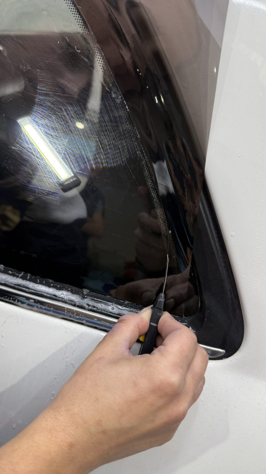 Découpe d’un film teinté au cutter sur une vitre latérale blanche, formation vitres teintées en Belgique